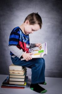 少年が沢山の本の上に座って、本を開いて読んでいます